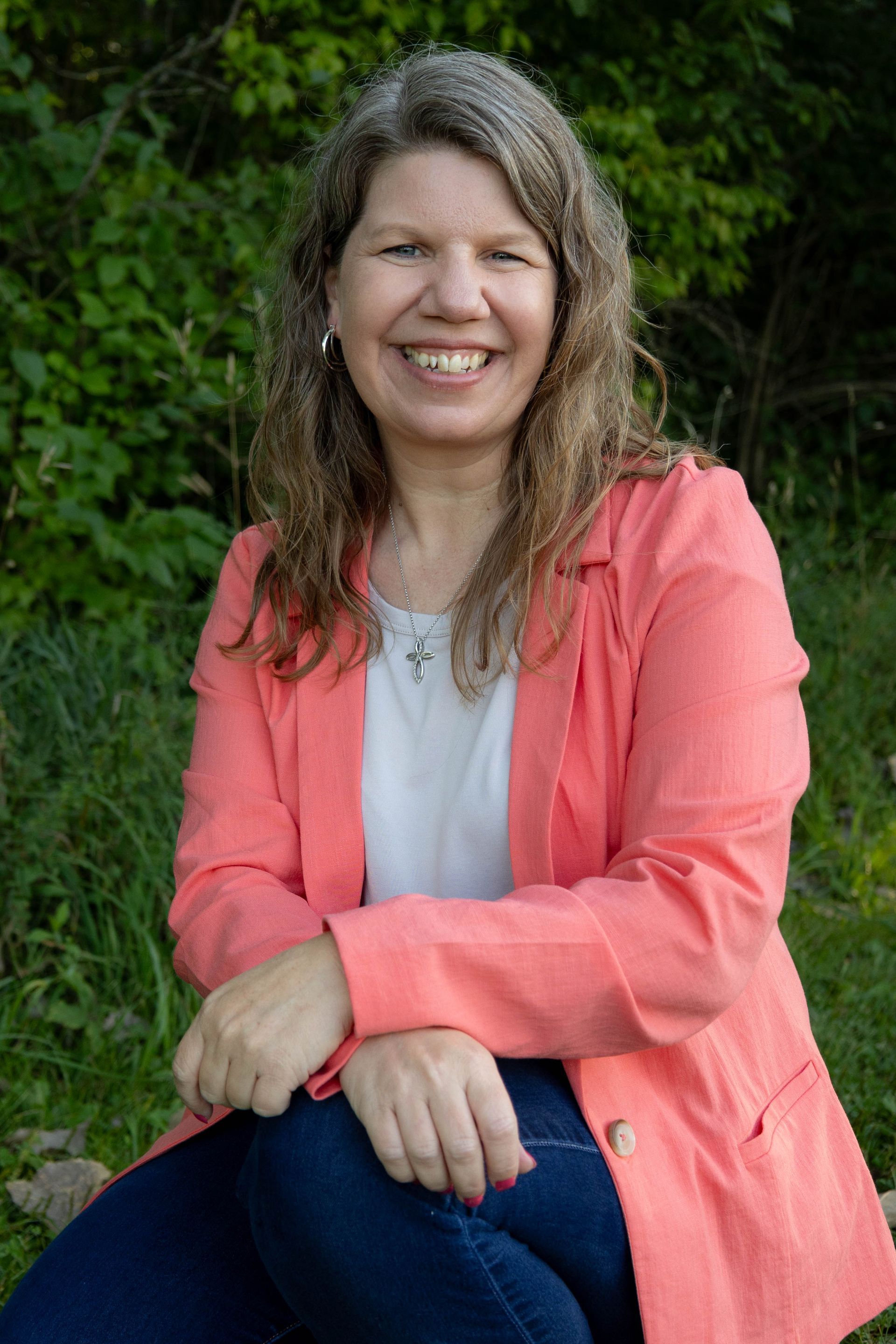 Woman with long, wavy gray hair, wearing a coral blazer, smiles outdoors.