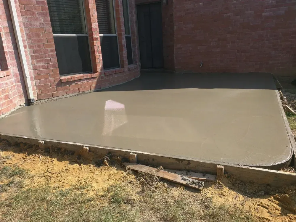 A freshly poured, wet concrete patio bordered by wooden forms against a brick house exterior. — Sand & Stone in Mudgeeraba, QLD