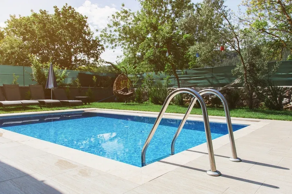A sunny backyard swimming pool with a metal ladder in the foreground, surrounded by green grass and trees. — Sand & Stone in Burleigh Heads, QLD