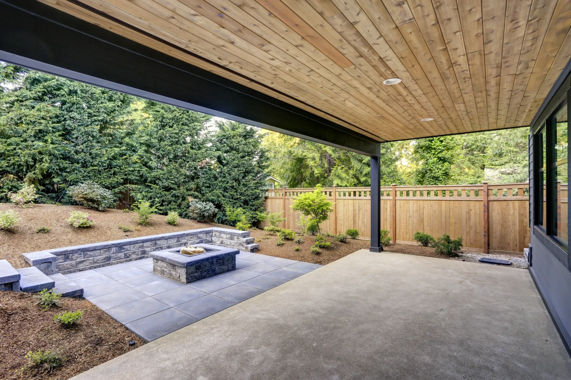 A covered patio with a concrete floor, a stone fire pit area, a low retaining wall, and a wood fence in a landscaped yard — Sand & Stone in Worongary, QLD