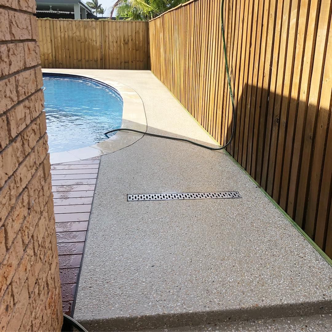 A concrete pool deck with a drainage grate next to a brick wall and wooden fence beside a swimming pool. — Sand & Stone in Mudgeeraba, QLD