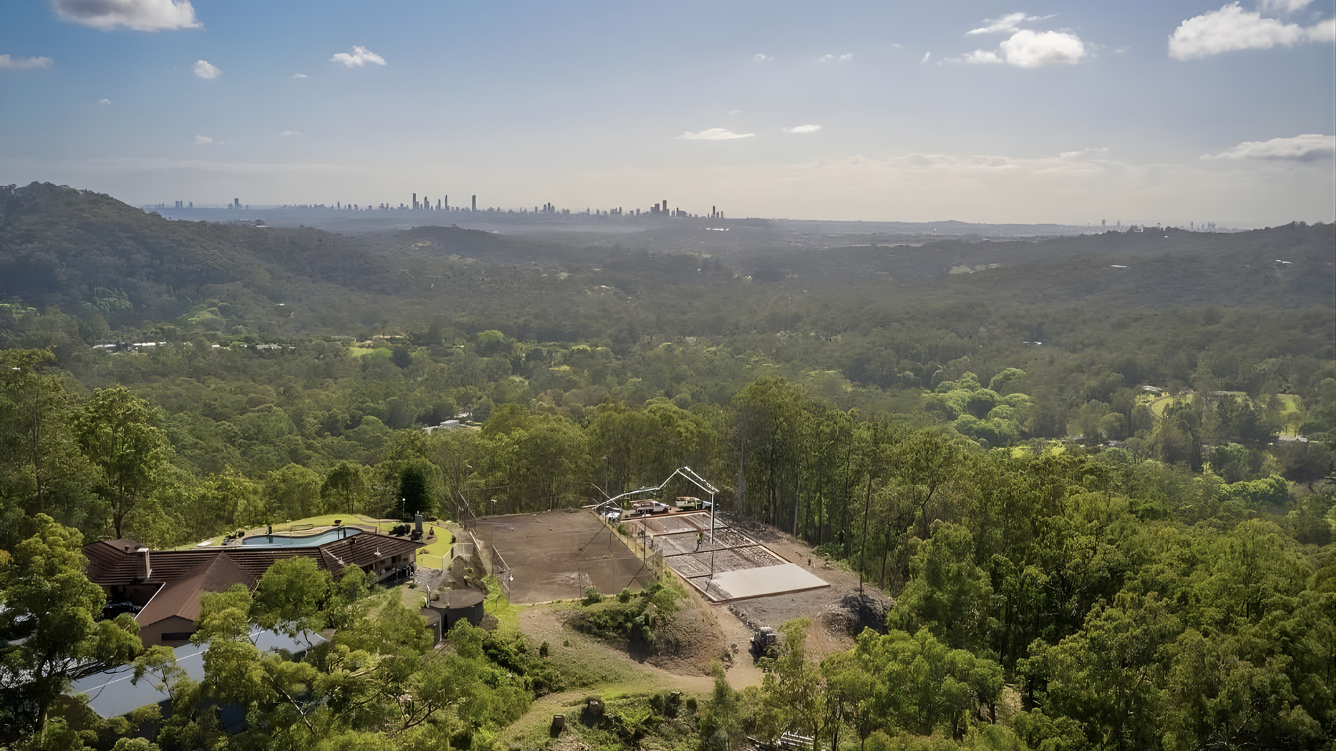 Wide green valley with rolling hills, scattered buildings, and a hazy city skyline in the distance — Sand & Stone in Worongary, QLD