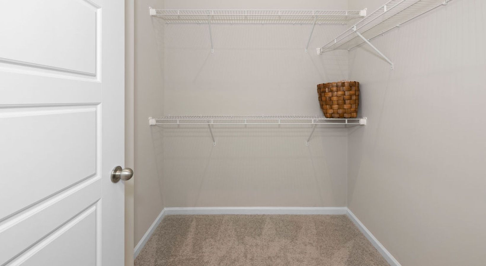 Empty walk-in closet with wire shelves, basket, beige carpet, and a partially open white door. We invite you to visit 1001 Tyvola and take a tour of our apartments with walk-in closets in Charlotte.
