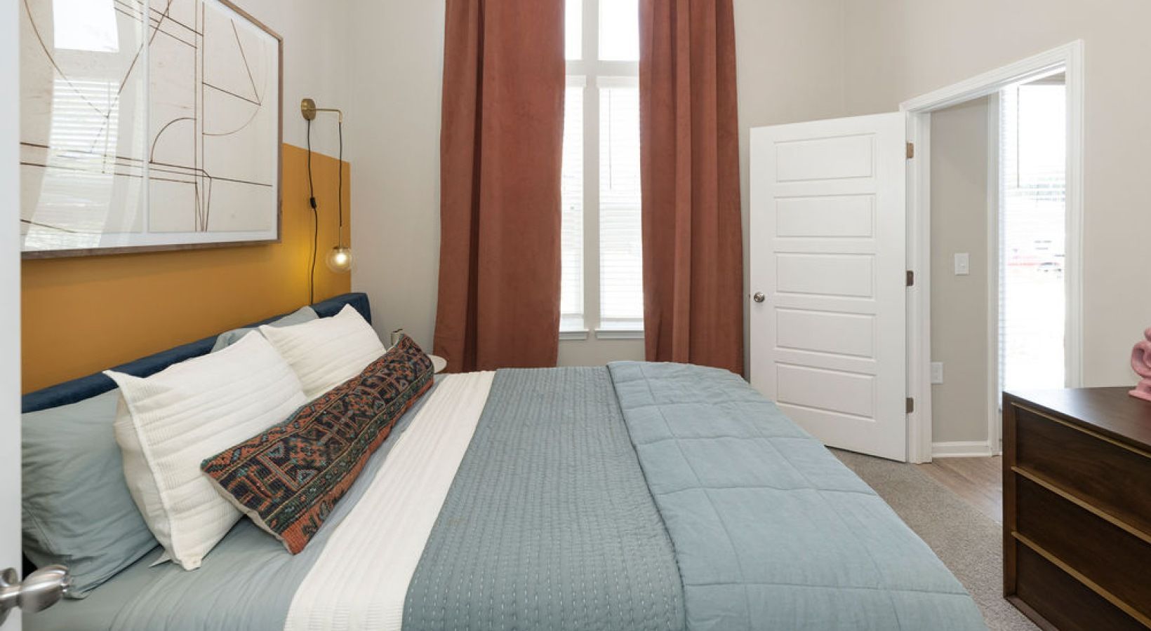 Bedroom with bed, orange wall accent, window with rust-colored curtains, door, and dresser. We invite you to visit 1001 Tyvola and take a tour of our apartments near Little Sugar Creek Greenway in Charlotte.