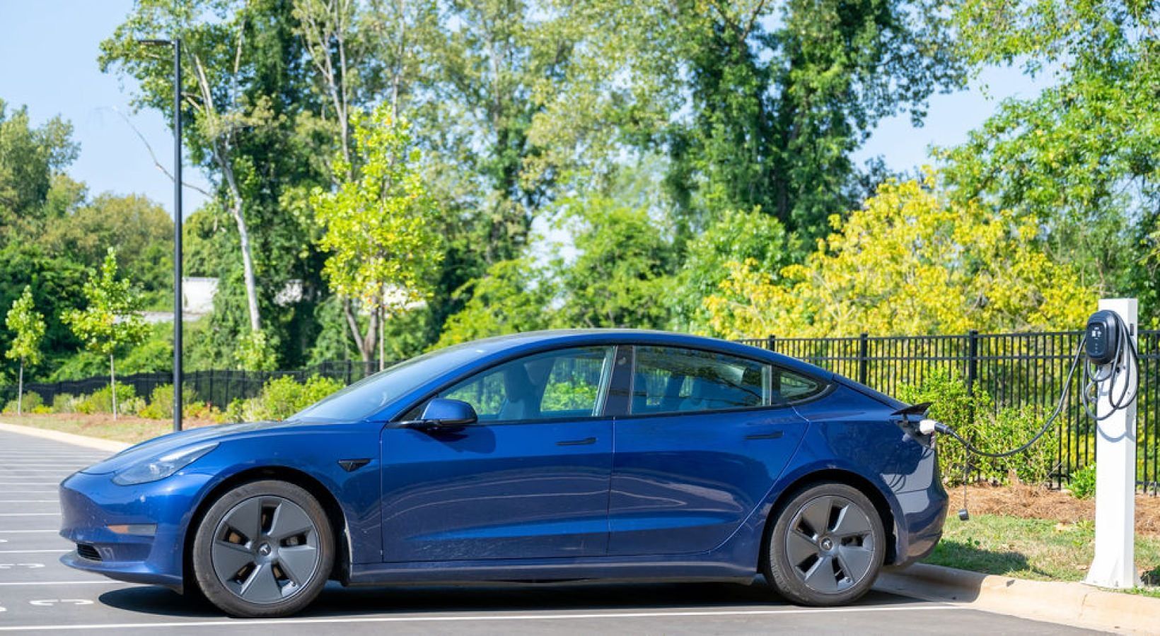 Blue Tesla sedan parked near a charging station in a parking lot, surrounded by trees.