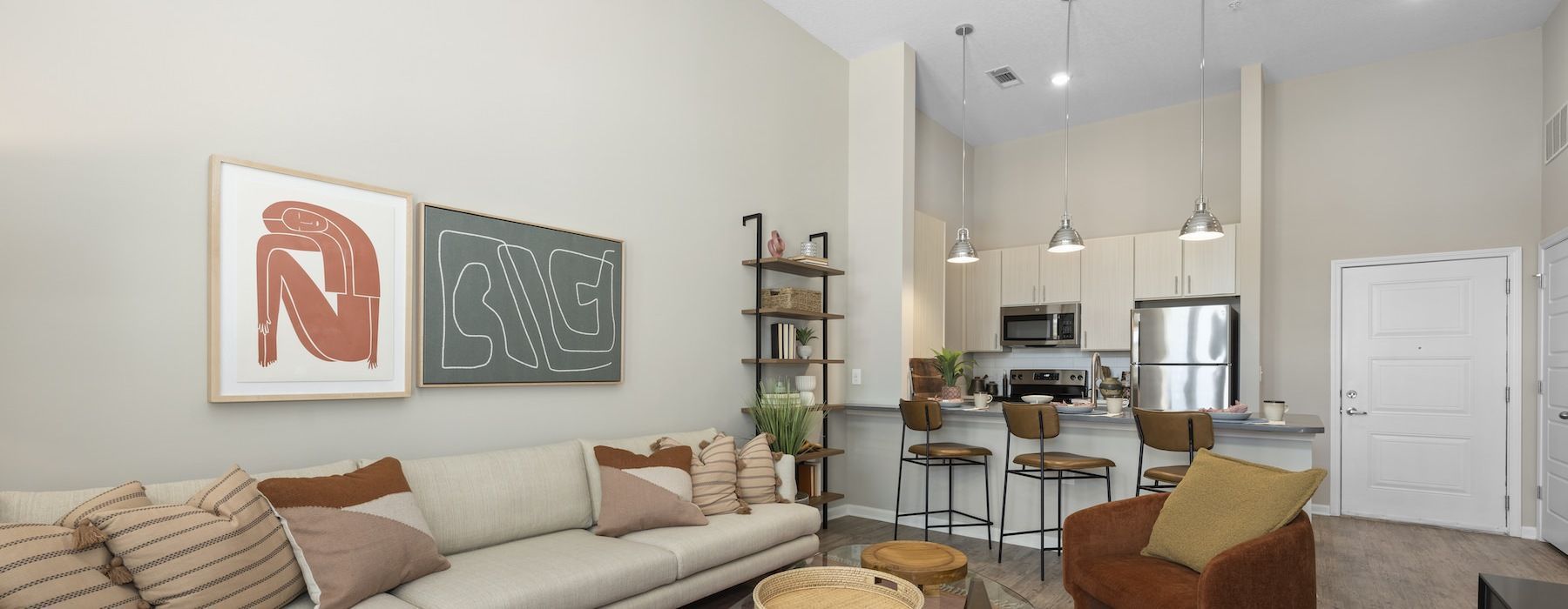 Living room with a sofa, kitchen, and decorative artwork. Neutral tones, high ceiling, natural light. We invite you to visit 1001 Tyvola and take a tour of our apartments near John Crosland School in Mecklenburg County.