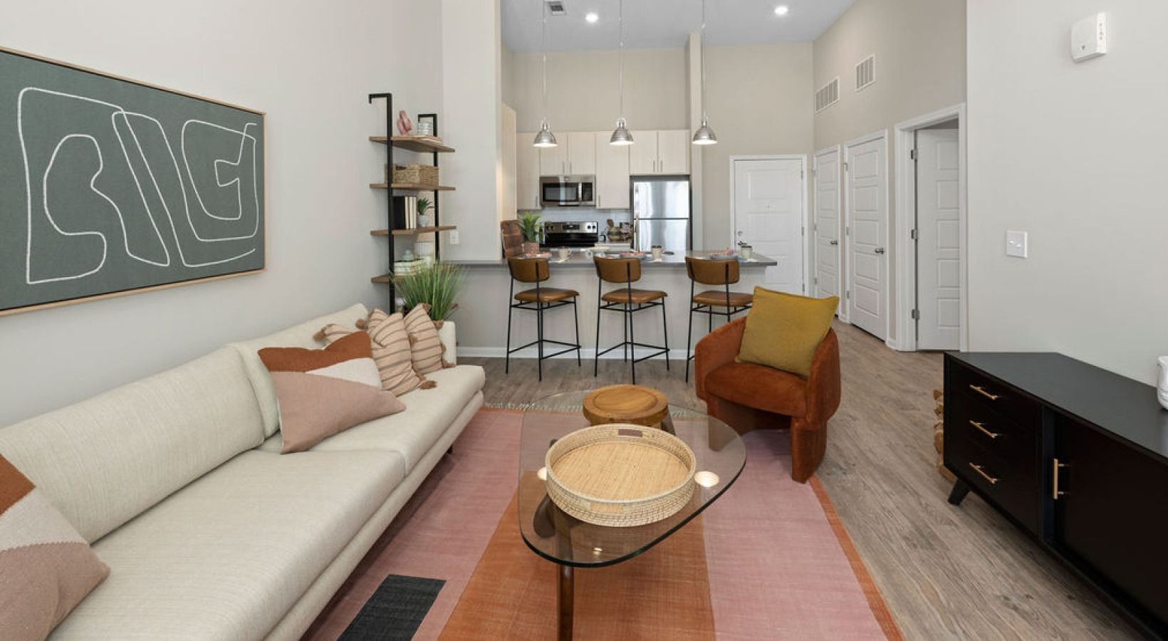 Living room with sofa, kitchen, and bar stools; neutral colors, rug, and artwork. We invite you to visit 1001 Tyvola and take a tour of our 3-bedroom apartments in Mecklenburg County.