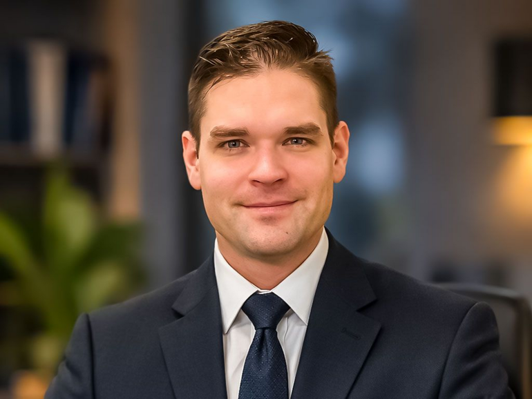 Portrait of a man in a dark suit and tie, smiling against a softly blurred indoor background
