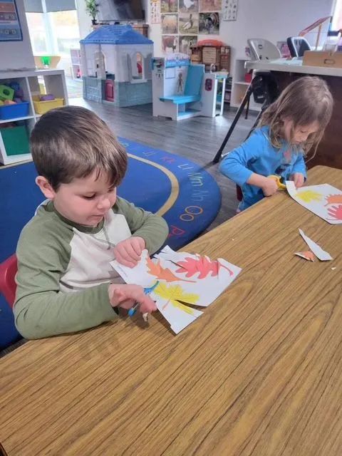 Two young children seated at a table, cutting paper with scissors at a daycare.