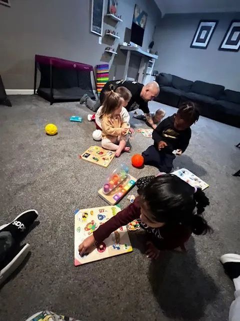 Children and a man playing with puzzles on a gray carpeted floor in a room.