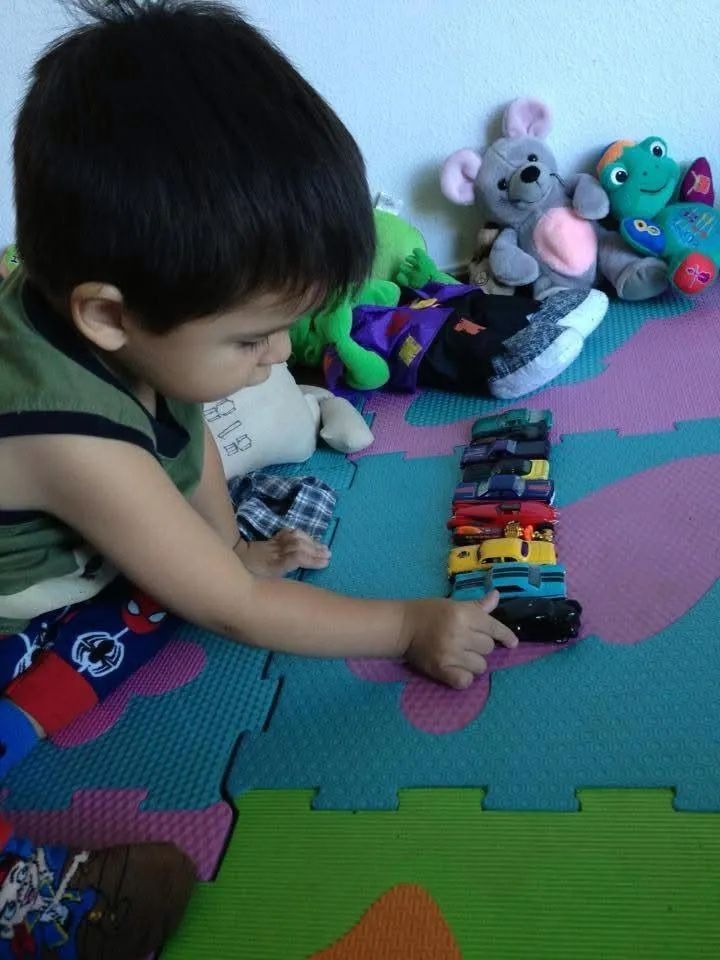 Young child stacking toy cars on a colorful floor. Toys and plush animals are in the background.