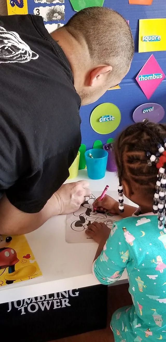 A man and child drawing at a table. The child is wearing pajamas. There are art supplies on the table.