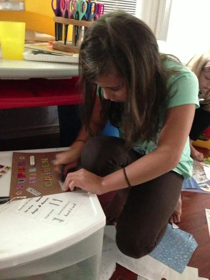 Girl kneeling, adding labels to a craft project, brown hair, green shirt, inside setting.