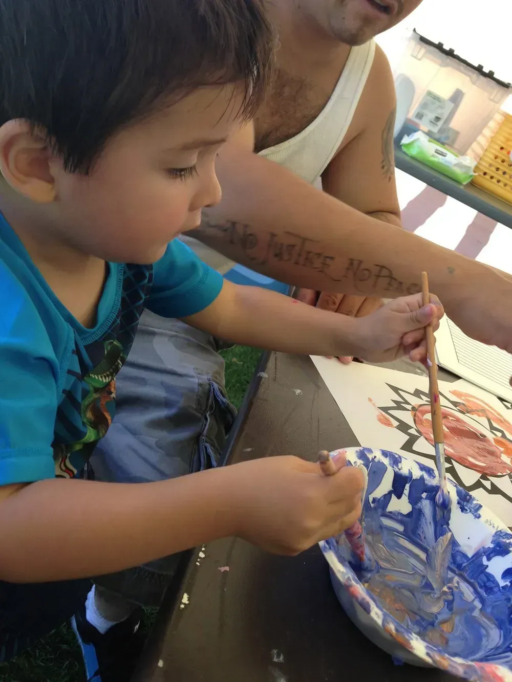 Young boy painting with blue paint, sitting at a table next to an adult in a sunny outdoor setting.