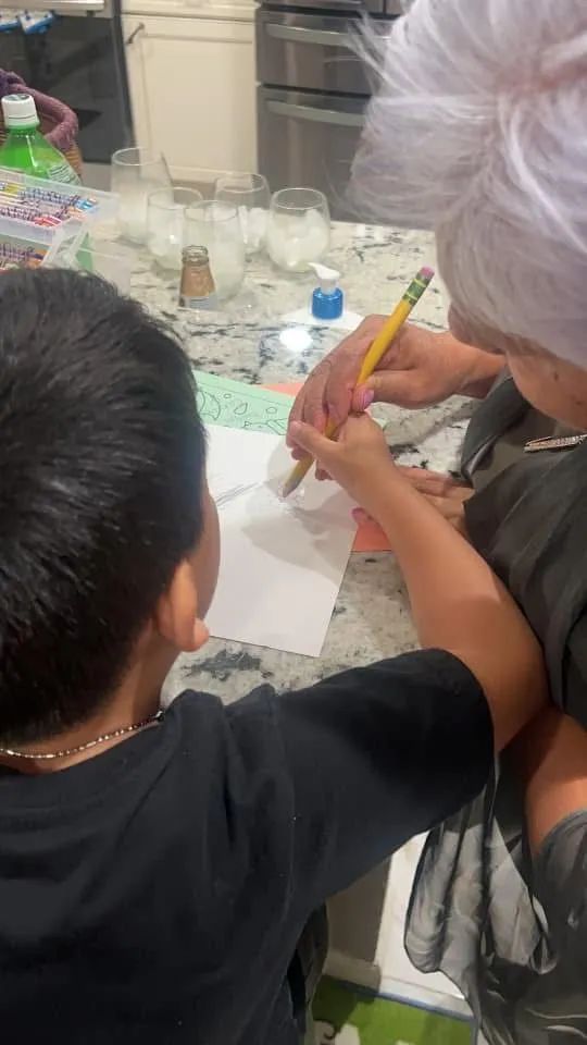 An elderly woman helps a young boy draw with a pencil on white paper in a kitchen.