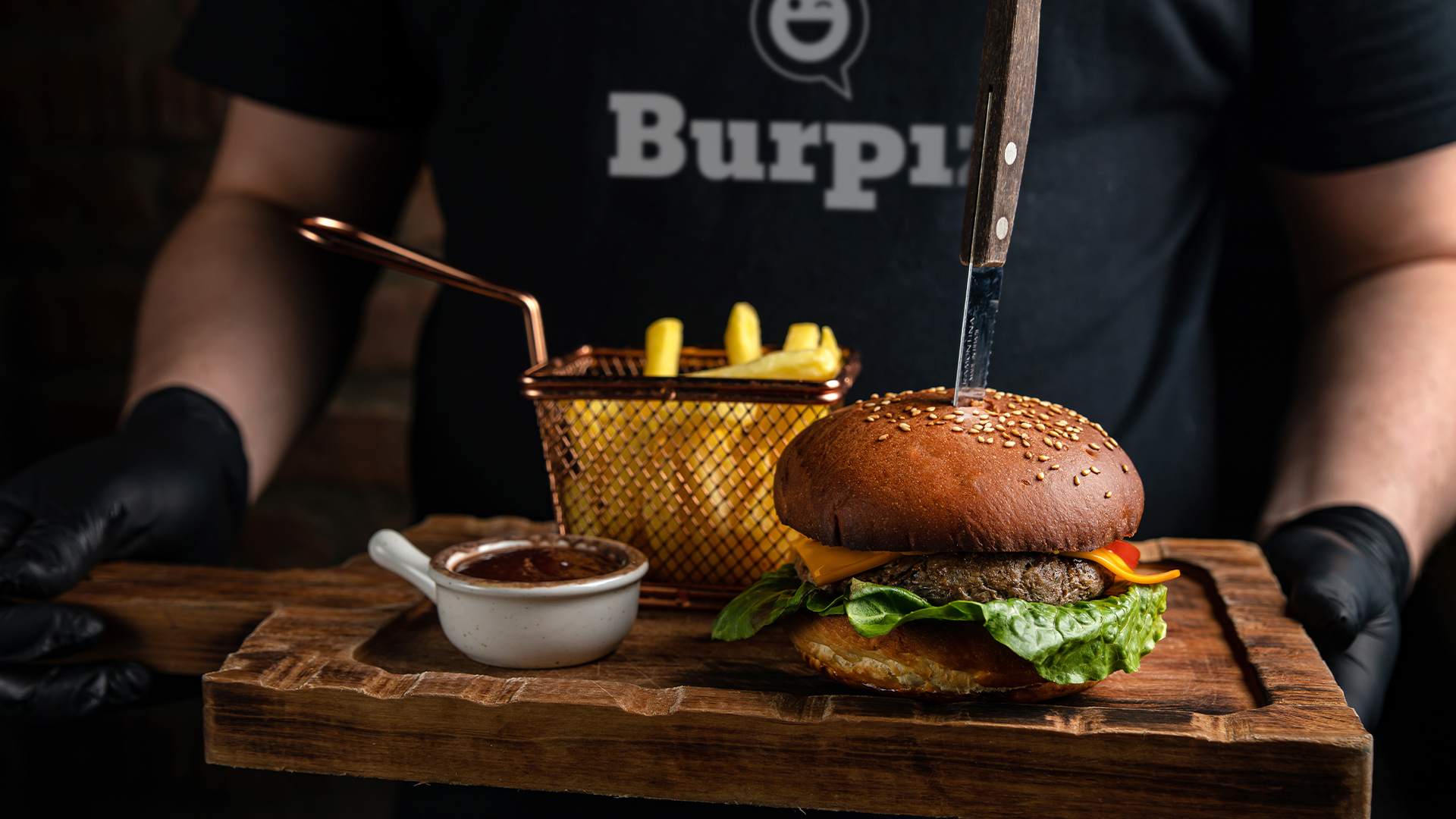 Pessoa segurando uma tábua de madeira com um hambúrguer, batatas fritas e molho. A pessoa usa luvas pretas e uma camiseta com a frase 