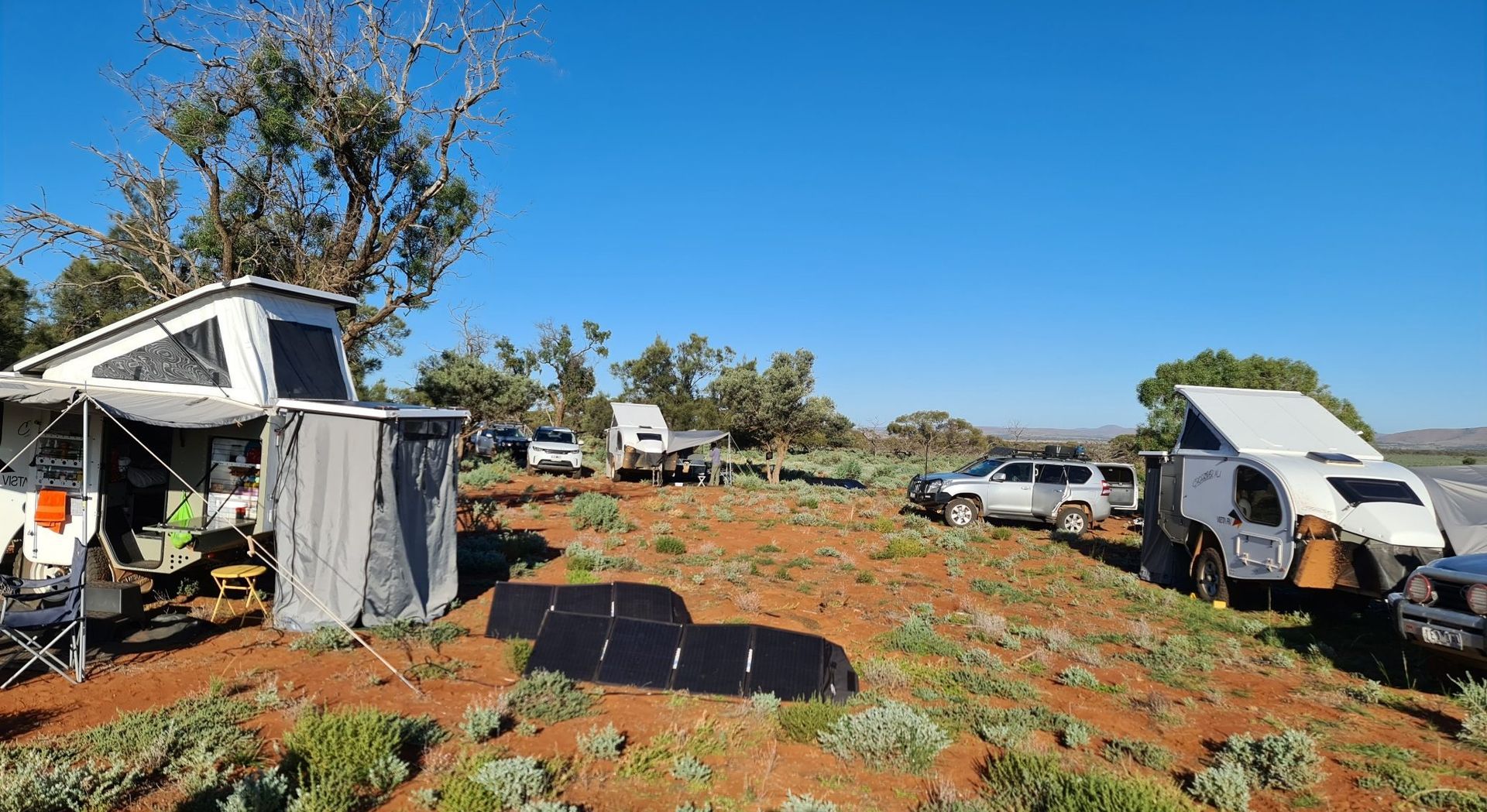 Two camping trailers and a vehicle parked on arid, red-dirt terrain under a bright blue sky.