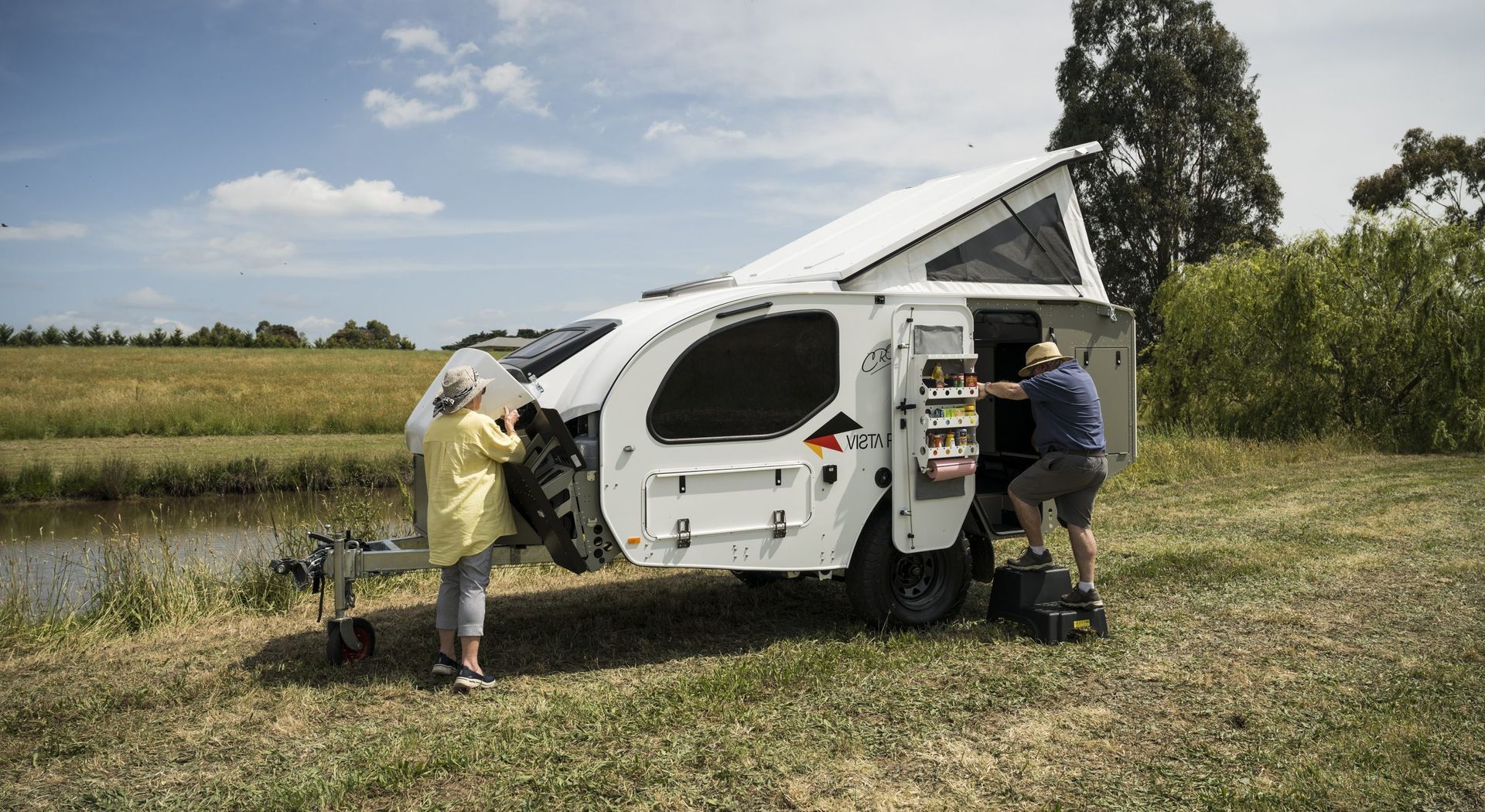 Two people set up a white pop-top camper trailer parked on grassy ground beside a pond under a partly cloudy sky.