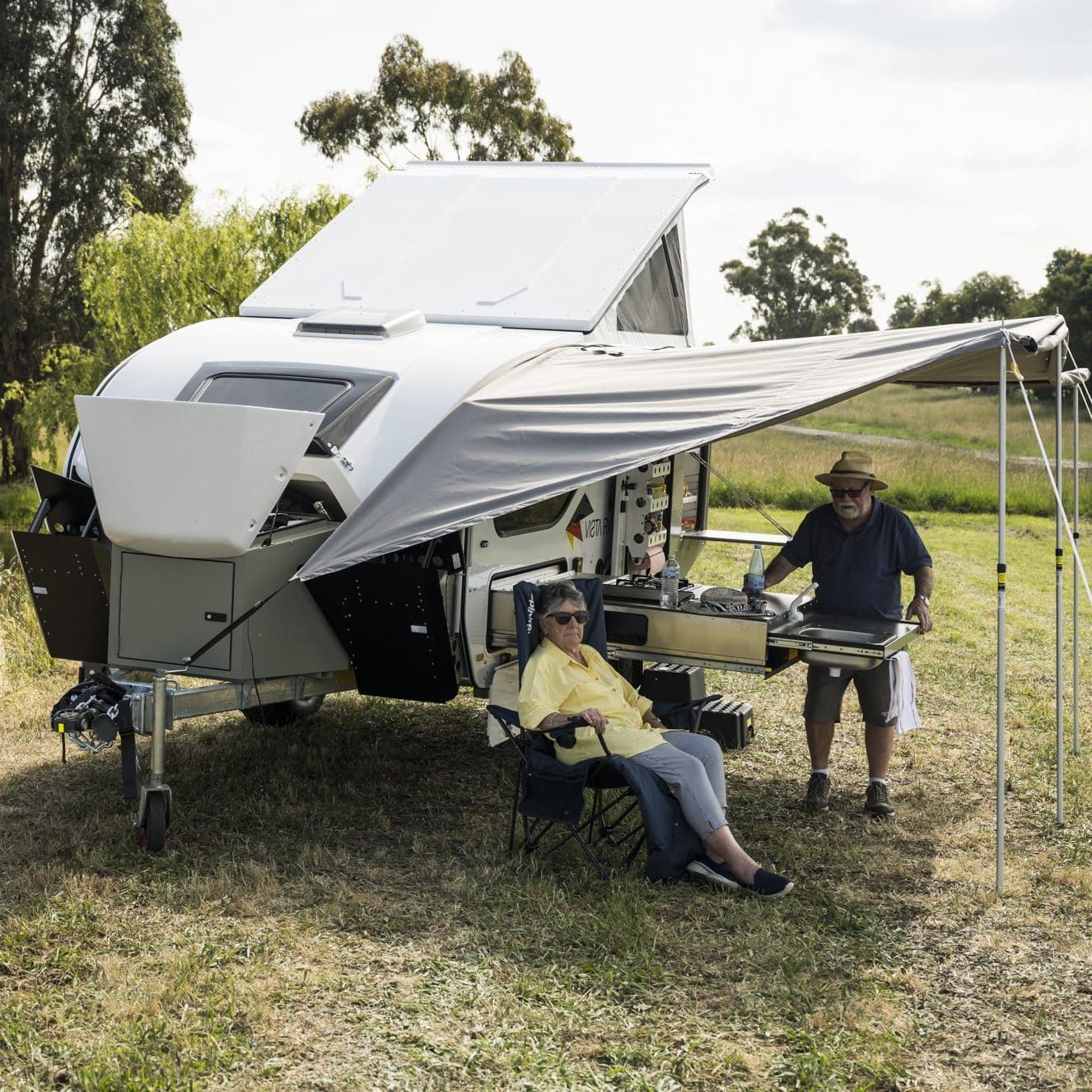 A camper trailer parked in a field with a deployed awning, a person seated in a chair, and another standing by a cooktop.