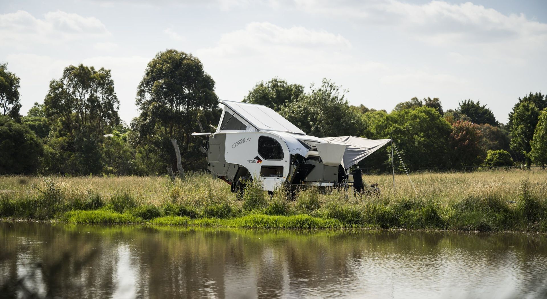 A modern, white and gray teardrop camper parked in a grassy field next to a calm lake, with an awning extended.