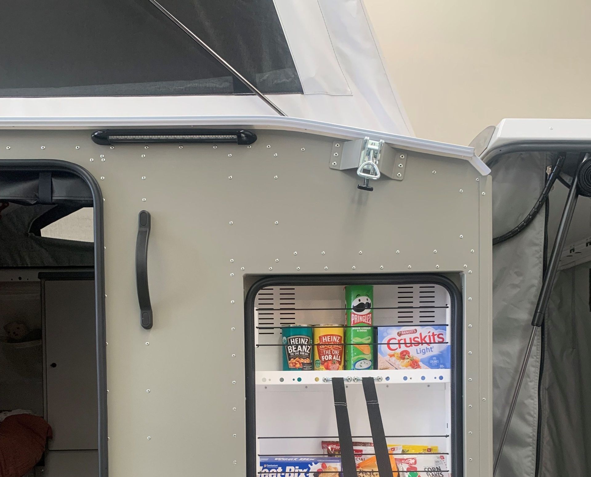 A side panel of a pop-up camper featuring an open exterior pantry cabinet stocked with various snack containers and boxes.