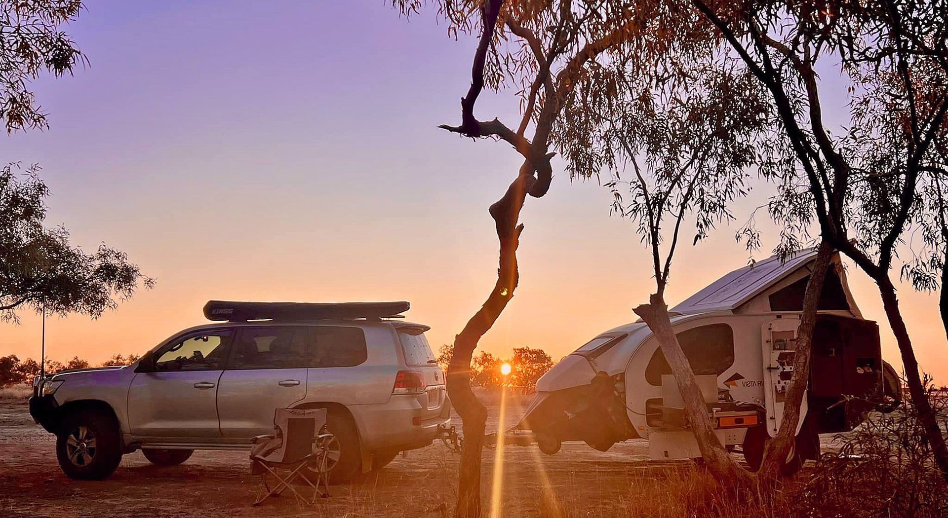 A silver SUV and a small teardrop camper parked in a wooded area under a sunset sky with a bright orange horizon.
