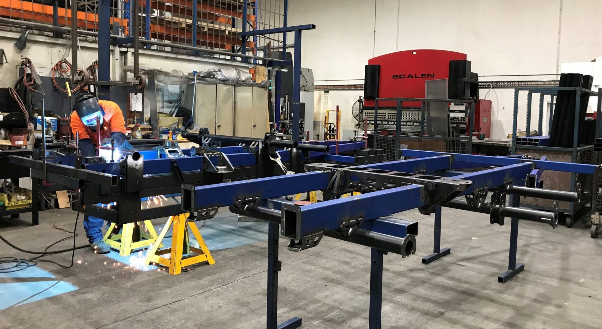 A person in high-visibility gear welding a blue metal frame in an industrial workshop.