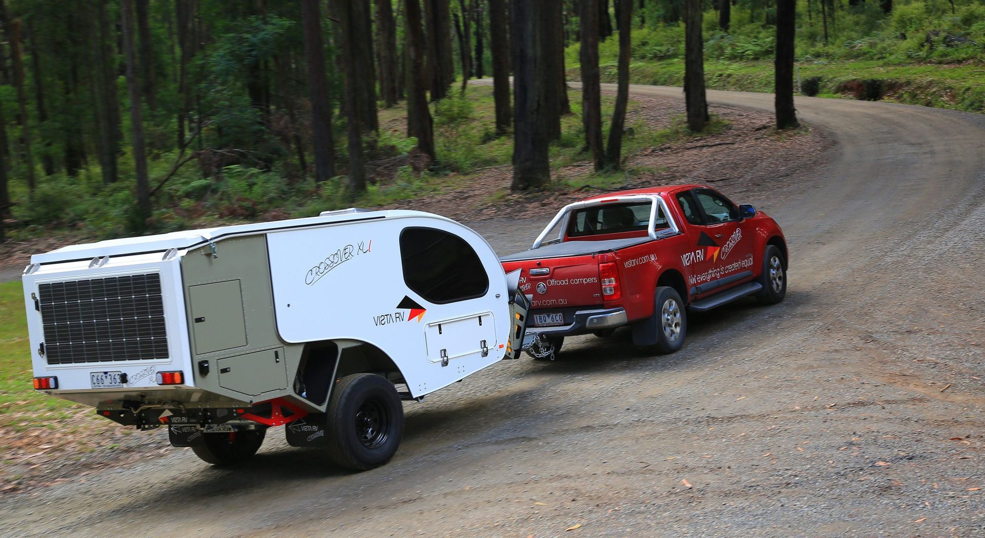 A red pickup truck tows a white off-road camper trailer along a dirt path through a forest.