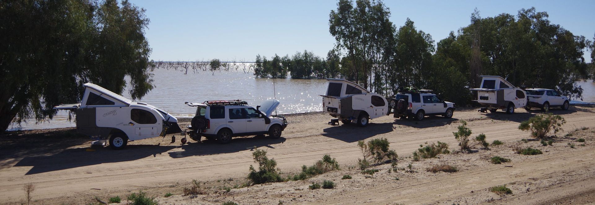 A line of white 4WD vehicles towing pop-top campers parked on a dirt track beside a large lake.