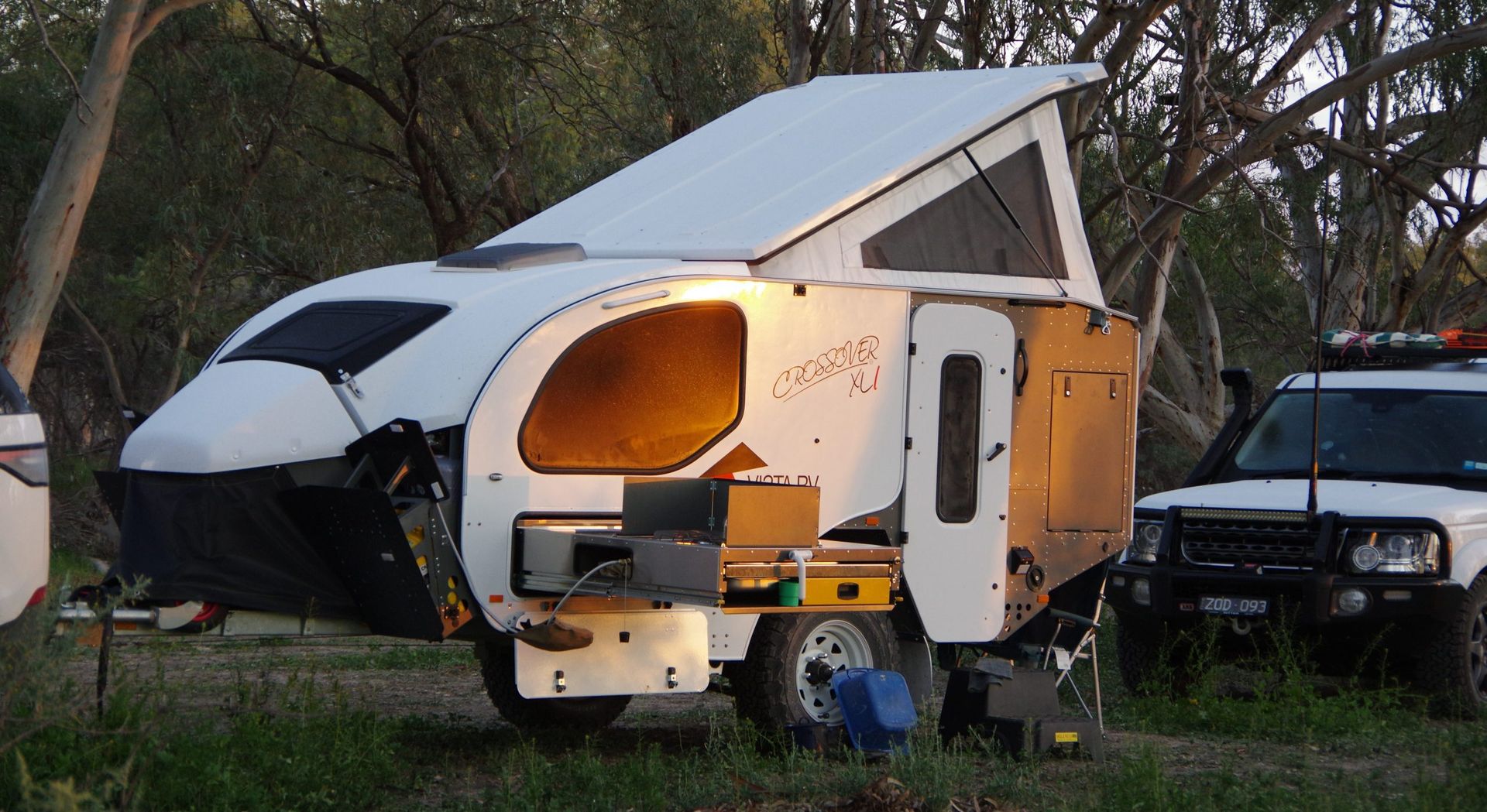 A white off-road camper trailer with a pop-top roof is parked in a wooded area next to a white SUV.