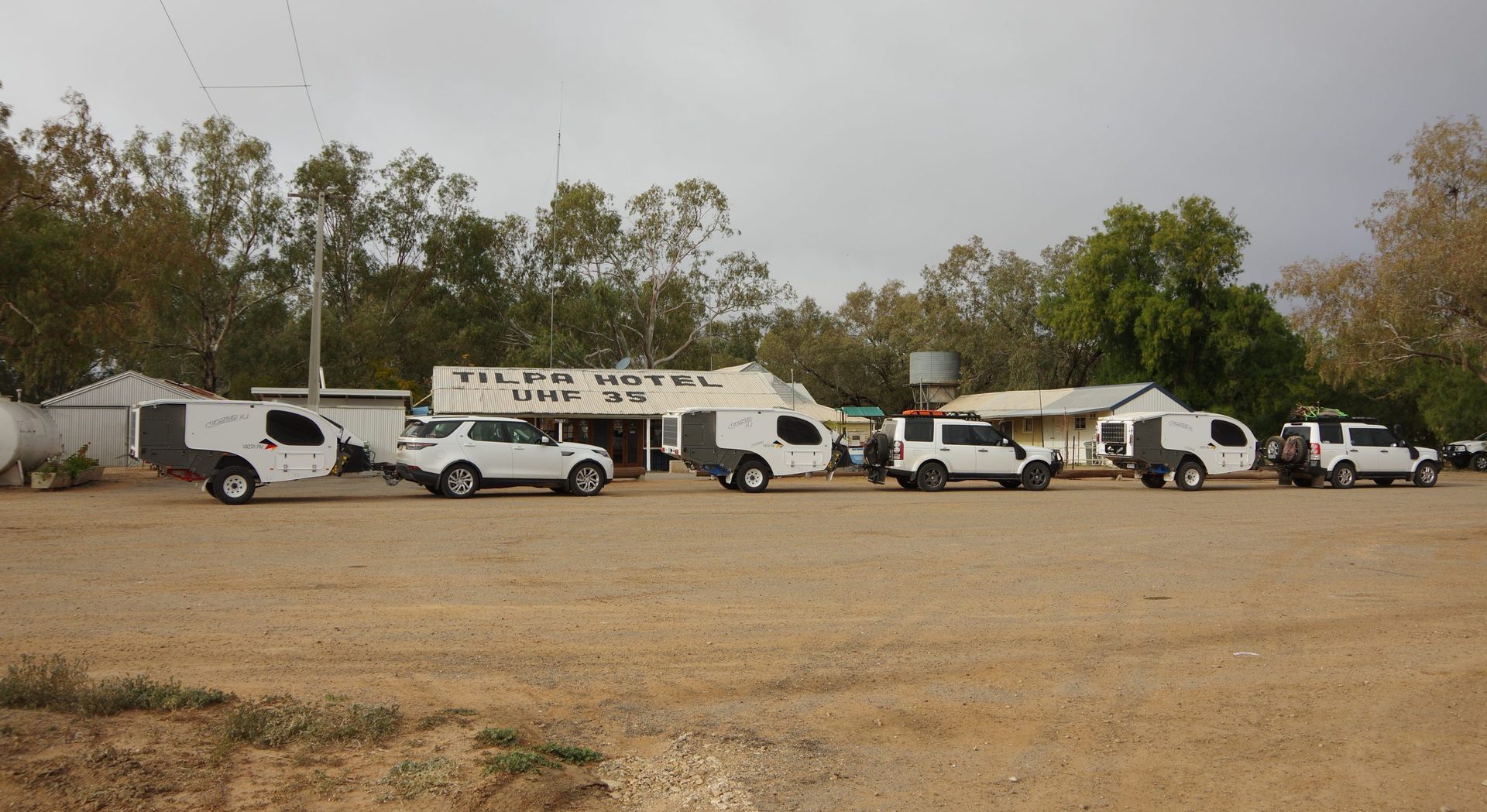 A line of white 4WD vehicles with matching camper trailers parked in front of the Tilpa Hotel under a cloudy sky.