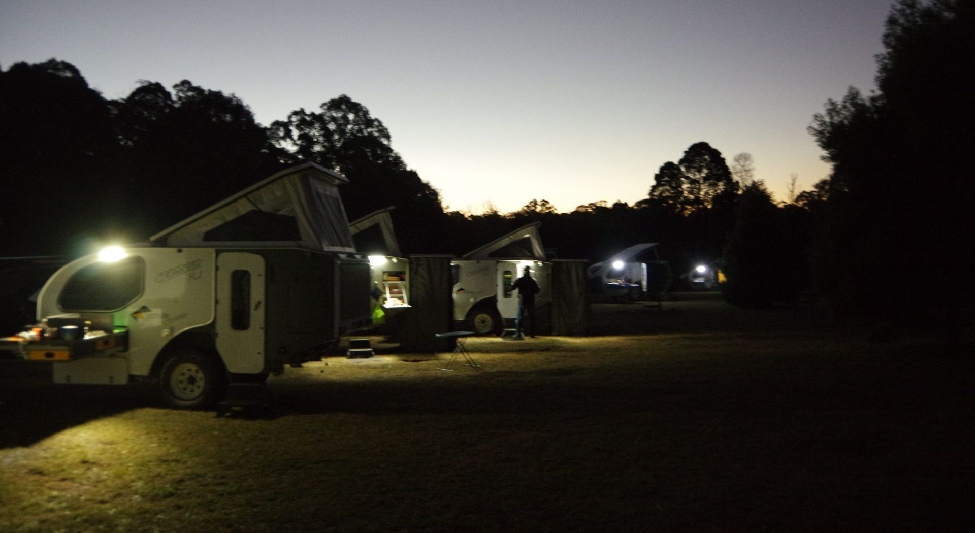 Three campers parked in a line at twilight, illuminated by their own exterior lights against a dark forest backdrop.