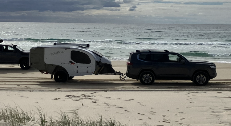 A dark SUV tows a white and gray teardrop-style camper along a sandy beach near the ocean.