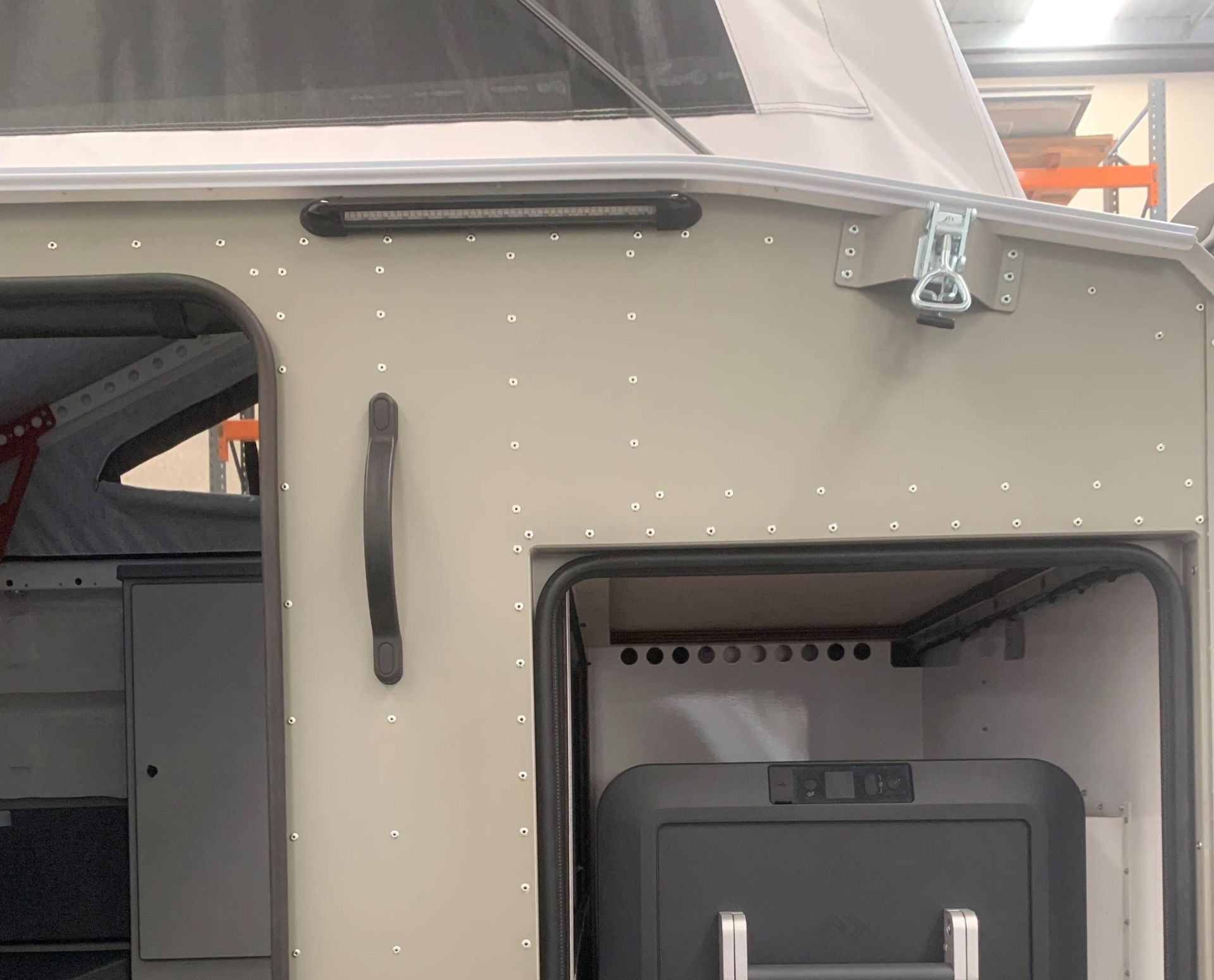 Close-up of a tan camper exterior with a side door, handle, black LED light, and a portable fridge inside the compartment.