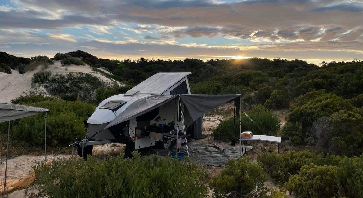 A camper trailer with an extended awning set up in a sandy, coastal landscape under a sunrise sky.