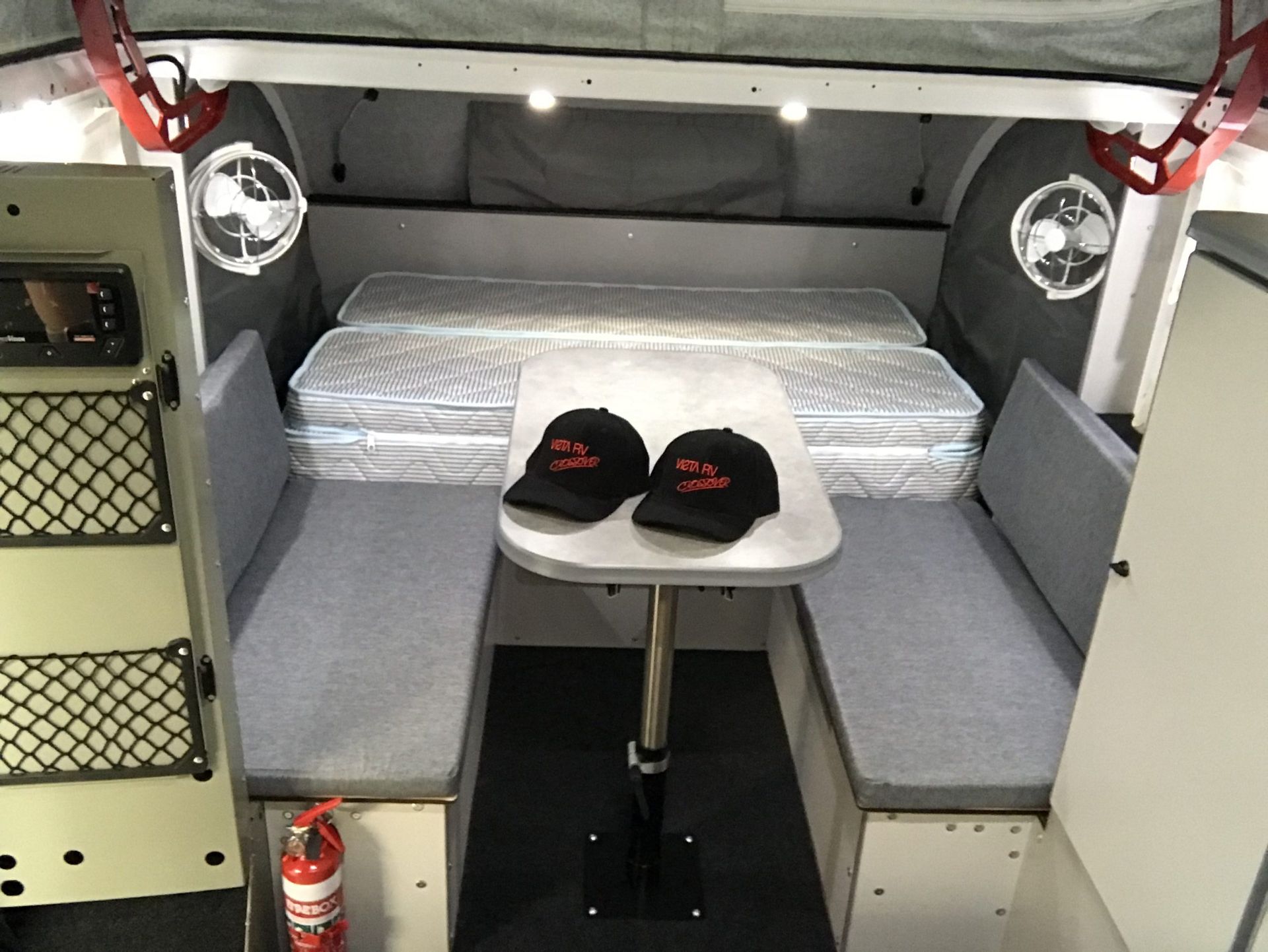 Interior of a camper featuring a dining area with two gray benches, a central table, two black caps, and sleeping mats.