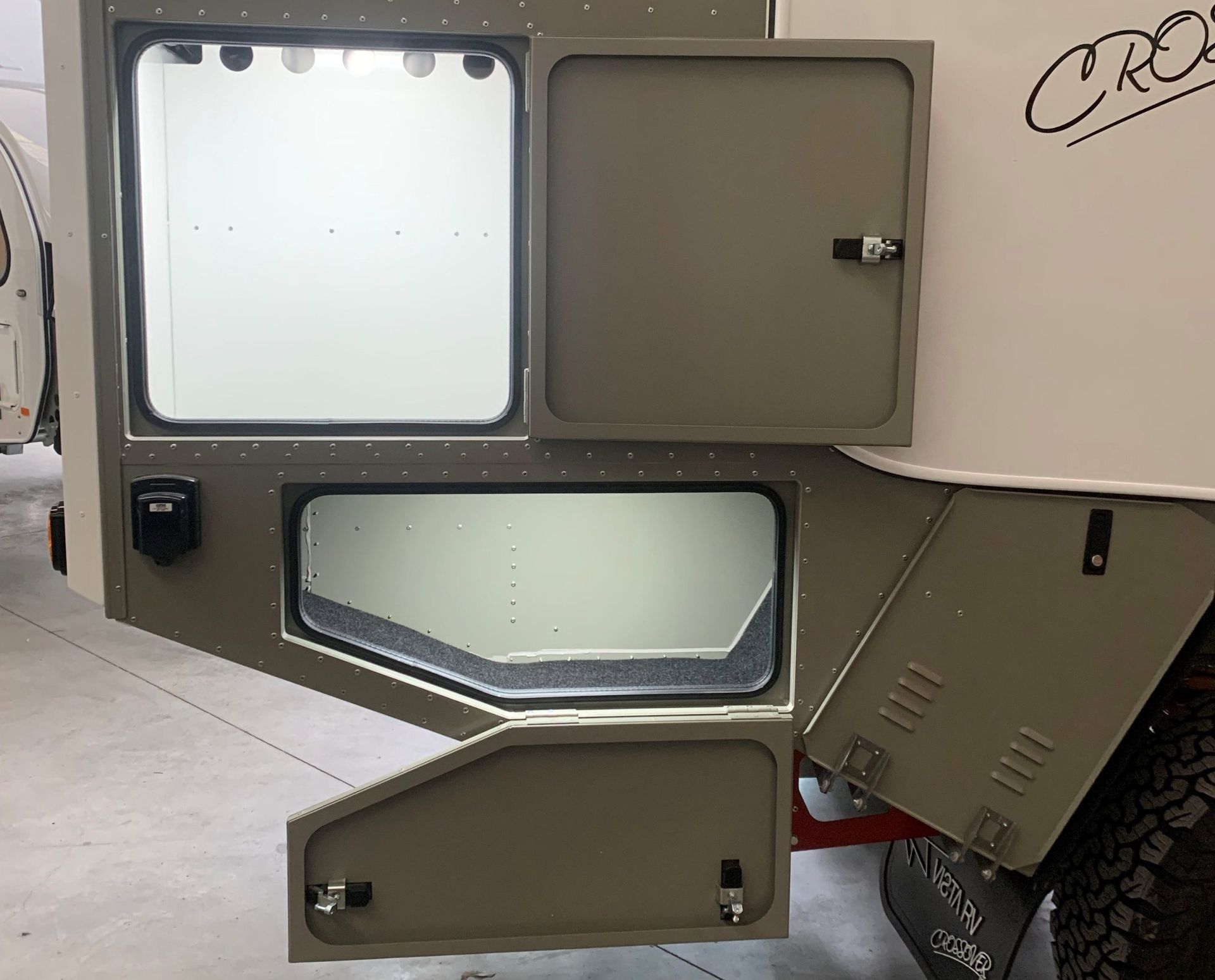 A side profile of an olive-green camper with two open storage compartments, one rectangular and one angled.