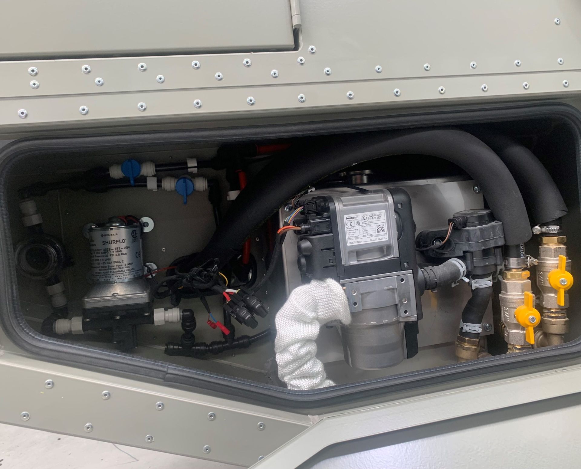 An open utility compartment on a vehicle exterior, revealing a water pump, heating unit, insulated hoses, and yellow valves.