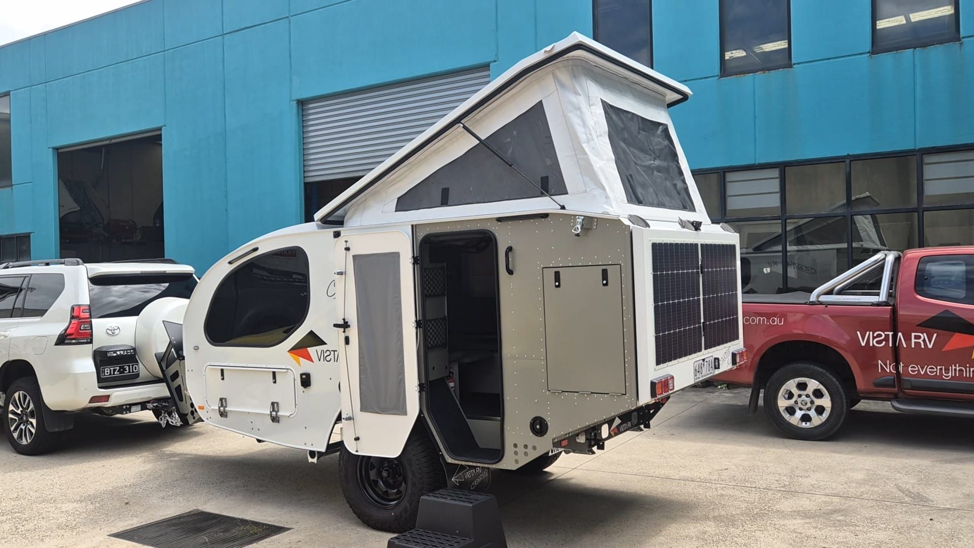 A small white camper trailer with a pop-up roof parked between a white SUV and a red pickup truck outside a blue building.