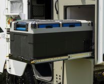 A portable, black and grey refrigerator pulled out on a metal slide rail from the back of an adventure vehicle.