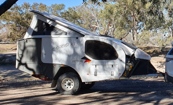 A white off-road camper trailer with a pop-top roof parked on dirt ground among trees in a sunny outdoor setting.