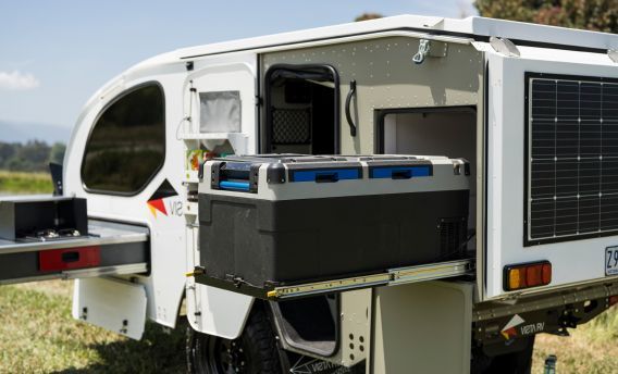 A white camper trailer in a field with a slide-out drawer holding a portable fridge and an outdoor kitchen area.