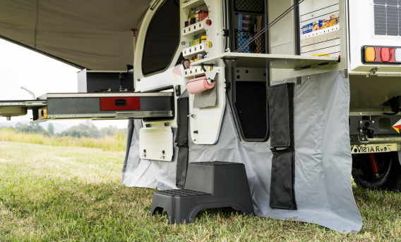 Side view of an off-road camper with an outdoor kitchen drawer extended, a step stool, and a gray fabric side skirt.