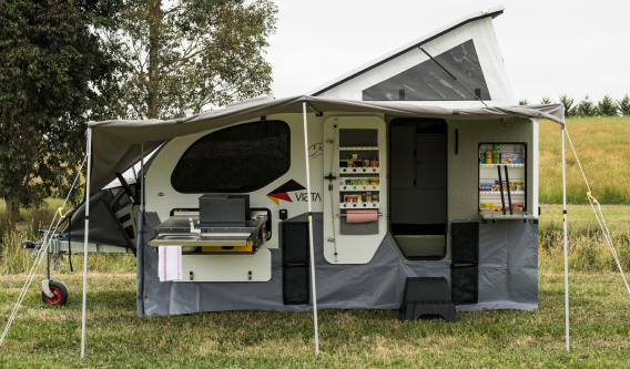 A white and grey pop-top camper trailer parked in a field, with an open side awning, outdoor kitchen, and pantry visible.