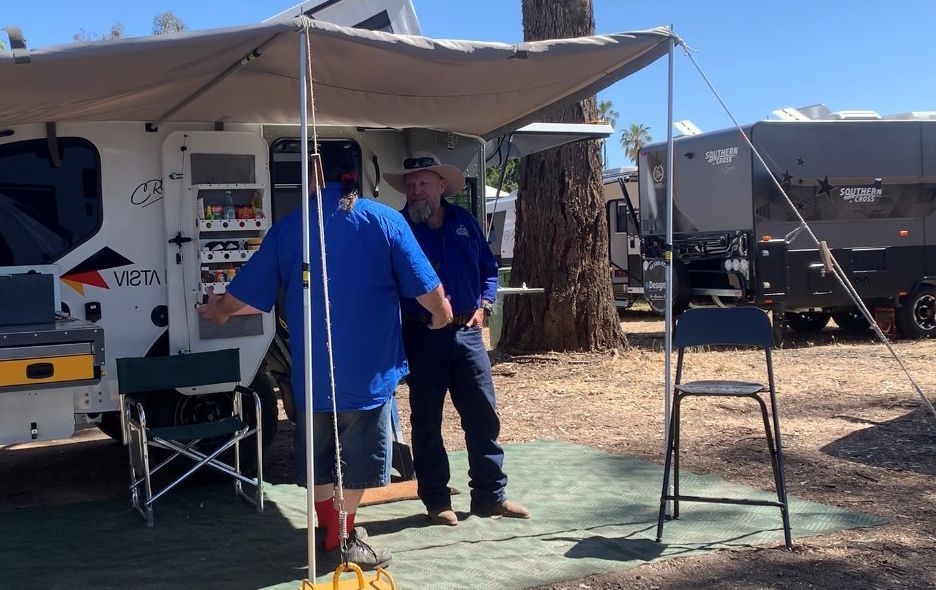 Two people in blue shirts stand talking under a camping trailer awning in a sunny, tree-lined outdoor campsite.