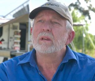 A person with a white beard and a baseball cap wearing a blue shirt, speaking in an outdoor setting.