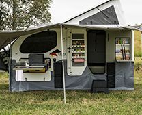 A popup camper set up on grass, featuring an outdoor kitchen, side shelves, and an awning extended for shade.