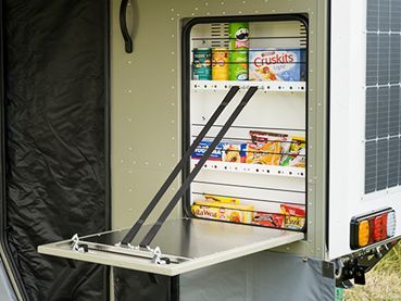 An open external pantry compartment on a camper trailer with shelves holding various food items.