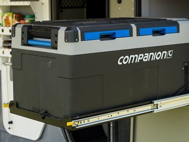 A gray and black Companion portable camping fridge pulled out on a sliding tray from a vehicle storage compartment.