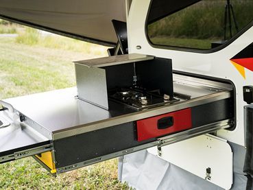 A stainless steel slide-out kitchen unit extends from the side of a camper, featuring a built-in stove and red drawer.